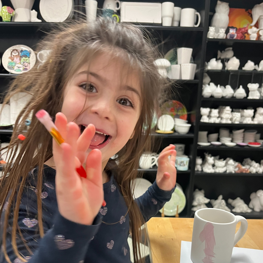 A happy child painting pottery in a bright studio, surrounded by shelves filled with unpainted ceramics.